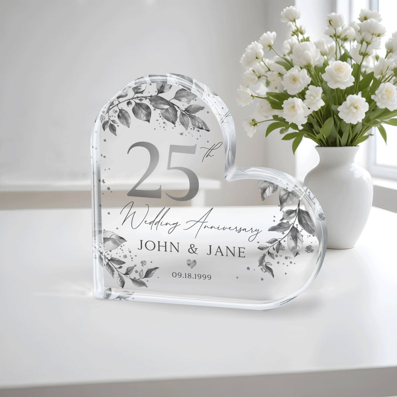 May include: A clear heart-shaped crystal paperweight with a silver design of leaves and the text "25th Wedding Anniversary John & Jane 09.18.1999".