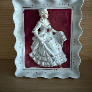 May include: A rectangular porcelain wall plaque with a scalloped edge and gold trim. The centre features a raised, three-dimensional white figurine of a person in a period gown against a deep red background, accented with gold floral details.