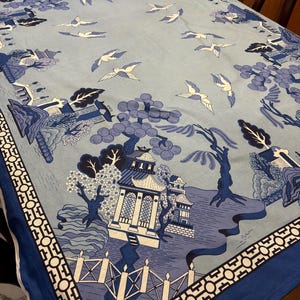 May include: A light blue tablecloth with an Asian-inspired design. The pattern features pagodas, trees, bridges, and flying birds in shades of blue and white. A dark blue border with a geometric pattern frames the edges.