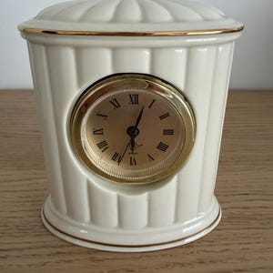 May include: A cream-coloured ceramic table clock with a fluted, cylindrical body and a matching lid. The clock face is gold with black Roman numerals and thin black hands, surrounded by a polished gold-tone circular frame and gold trim.