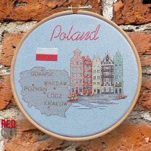 Könnte beinhalten: Ein Kreuzstich-Stickrahmen mit dem Wort "Poland" in roter Schrift. Das Design umfasst die polnische Flagge, eine Karte von Polen mit Städtenamen und farbenfrohen Gebäuden. Der Rahmen ist vor einer Backsteinmauer angebracht.