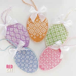 May include: Five colorful, cross-stitched Easter egg ornaments. Each egg features a unique geometric pattern in shades of purple, orange, green, blue, and pink. They are adorned with white satin bows and hanging loops. The logo "RED CATE" is visible.
