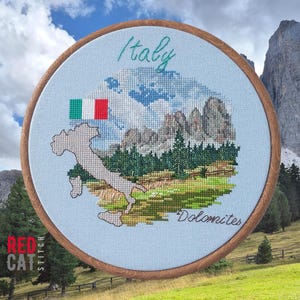 May include: Cross-stitch embroidery of Italy's Dolomites mountains, featuring the Italian flag and a map outline. The design is set within a wooden embroidery hoop, with the word "Italy" and "Dolomites" stitched in green.