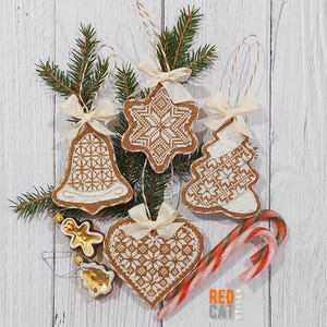 Peut inclure: Ornements de Noël brodés en forme de cloche, étoile, sapin et cœur, avec des motifs au point de croix. Chaque ornement est orné d'un nœud crème et d'une boucle en ficelle. Deux cannes en sucre et des petits bonhommes en pain d'épices sont également présents.