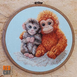 May include: Cross-stitch embroidery of two monkeys, one gray and one orange, hugging on a light blue background. The embroidery is set in a round wooden hoop. The text "RED CAT" is in the lower left corner.