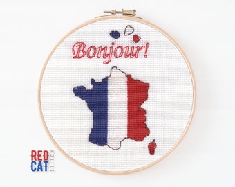 May include: A white embroidery hoop with a cross-stitch design of the French flag in the shape of France. The text "Bonjour!" is stitched in red above the flag.