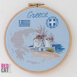May include: Cross-stitch embroidery in a wooden hoop featuring a light blue background with the word "Greece" and a map outline. The design includes Greek flag, a cross, and a depiction of windmills.