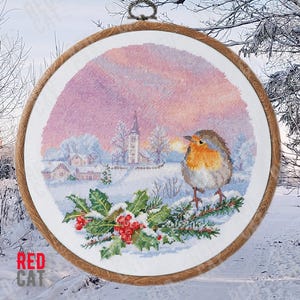 Op de afbeelding: Een borduurring met een wintertafereel in kruissteek. De scène toont een klein dorp met een kerk, een roodborstje dat op een hulsttak zit en een besneeuwd landschap met een roze en paarse zonsondergang.