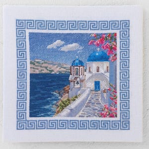May include: Cross-stitch of a Greek Orthodox church with blue domes and a white facade, set against a blue sea and sky. The image is framed by a blue Greek key border. Pink flowers and a coastal landscape complete the scene.