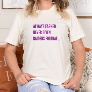 May include: A white t-shirt with purple text that reads "ALWAYS EARNED. NEVER GIVEN. RAIDERS FOOTBALL."
