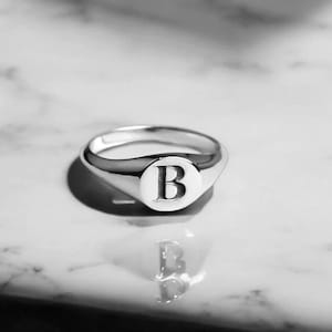 Puede incluir: Un anillo de sello plateado con la letra "B" grabada en una cara circular. El anillo está muy pulido y refleja la luz, sobre una superficie de mármol. Una pieza de joyería clásica.