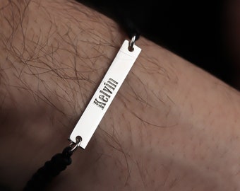 Op de afbeelding: Een zilveren armband met een zwart koord en een rechthoekig label met de naam "Kelvin" gegraveerd.