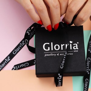 Op de afbeelding: Een zwarte geschenkdoos met een wit lint en de tekst "Glorria jewellery & accessories sense" erop gedrukt. Het lint is gebonden in een strik en de doos is open.