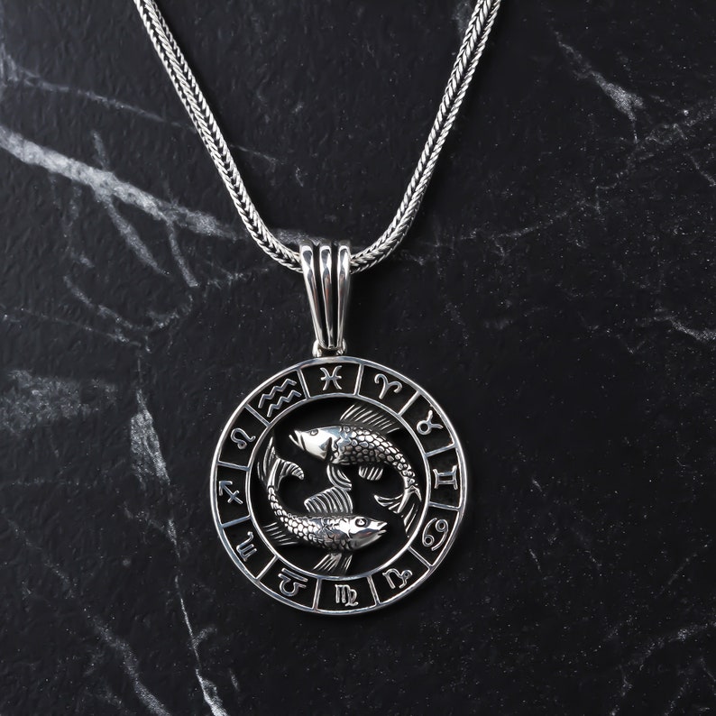 Op de afbeelding: Zilveren ketting met een ronde hanger met het sterrenbeeld Vissen, twee in elkaar verweven vissen, omringd door de astrologische tekens van de dierenriem.
