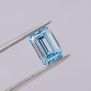 May include: A rectangular, light blue gemstone is held by silver tweezers. The gem features a step-cut design, with visible facets and internal reflections. The background is a soft, neutral grey.