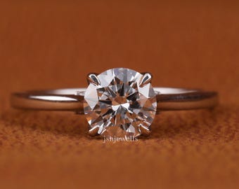 Könnte beinhalten: Ein Solitär-Verlobungsring mit einem rund geschliffenen Diamanten, der in einem dünnen, silbernen Band gefasst ist. Der Diamant funkelt hell vor einem warmbraunen Hintergrund. Der Text "jshjewells" ist auf dem Ring zu sehen.