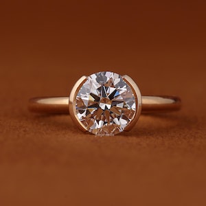 Può includere: Un anello d'oro con un grande diamante rotondo e trasparente incastonato in una lunetta. L'anello è su uno sfondo marrone e il diamante brilla. La fascia dell'anello è sottile e dorata.