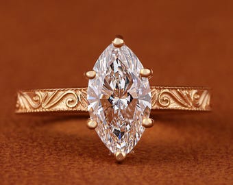 May include: A gold marquise-cut diamond ring. The diamond is set in a gold band with an ornate, engraved design. The ring's band features a detailed, swirling pattern. The ring is set against a brown background.