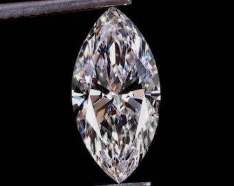 May include: A marquise-cut diamond held by tweezers against a black background. The diamond is clear and faceted, reflecting light to create a brilliant sparkle. The elongated shape is symmetrical with pointed ends.