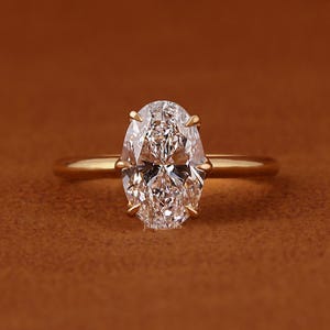 Puede incluir: Un anillo de oro con un diamante de talla ovalada. El diamante está engastado con cuatro garras, y la banda es de oro liso. El anillo se muestra sobre un fondo marrón.