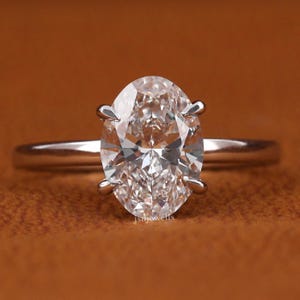 Può includere: Primo piano di un anello con diamante con un diamante taglio ovale. Il diamante è incastonato su una sottile fascia d'argento. L'anello è presentato su uno sfondo marrone. Il diamante è il punto focale, mostrando la sua brillantezza.