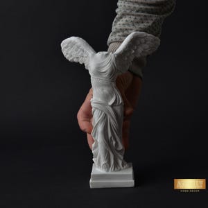 Puede incluir: Estatua de mármol blanco de la Victoria Alada de Samotracia, una diosa griega de la victoria, con alas extendidas. La estatua está sobre una base blanca y está siendo sostenida por una mano.