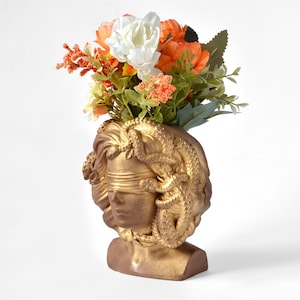 Puede incluir: Maceta de busto de color dorado con un rostro vendado y serpientes alrededor de la cabeza, sosteniendo un ramo de flores blancas y naranjas.