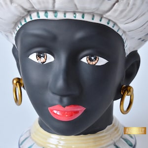 Könnte beinhalten: Eine Keramikkopfskulptur mit schwarzem Gesicht, roten Lippen und goldenen Creolen. Die Skulptur hat eine weiße Kopfbedeckung mit grünen Akzenten und einen gelb-blauen Kragen. Die Augen sind mit Braun und Weiß detailliert.