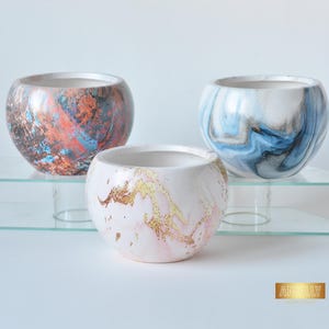 Può includere: Tre vasi di ceramica rotondi decorativi con disegni marmorizzati. Uno presenta un mix di arancione, blu e marrone. Un altro ha vortici blu e bianchi, e il terzo ha marmorizzazione rosa e oro. Sono su un espositore di vetro trasparente.