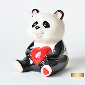 Peut inclure: Figurine de panda en céramique, principalement blanche avec des détails noirs, tenant un cœur rouge brillant. Le panda a des yeux, des oreilles et des membres noirs, avec des accents roses sur les oreilles et les pieds. Objet décoratif.