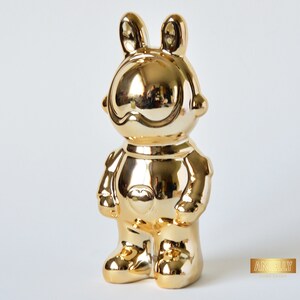May include: A gold-colored ceramic figurine of a rabbit wearing a space suit. The figurine has a large, round head with a wide smile and large, round eyes. The rabbit is standing with its arms at its sides and its legs slightly apart.