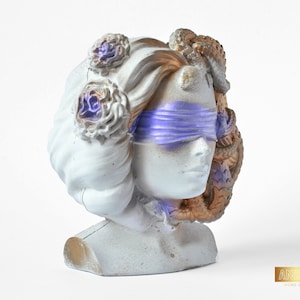 Könnte beinhalten: Eine weiße Gipsbüste einer Frau mit Augenbinde, goldenen Schlangen und lila Blumen. Die Büste ist ein dekoratives Wohnaccessoire.