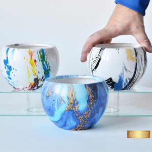 Könnte beinhalten: Drei dekorative Keramikschalen mit einzigartigen abstrakten Designs. Eine Schale hat blaue, gelbe und rote Farbspritzer, eine andere blaue und goldene Wirbel, und die dritte hat schwarze und goldene Pinselstriche. Die Schalen werden auf einem Glasregal ausgestellt.