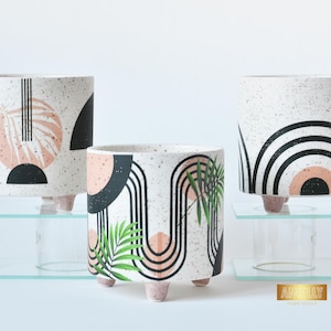 Puede incluir: Tres macetas decorativas de cerámica con diseños de terrazo. Las macetas presentan diseños geométricos en negro, melocotón y verde, con un fondo blanco moteado. Cada maceta tiene pequeños pies de color rosa claro.