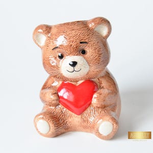 Puede incluir: Figura de oso de peluche de cerámica marrón con un corazón rojo. El oso tiene una cara sonriente, ojos negros y detalles blancos en las patas y las orejas. Es un artículo decorativo.