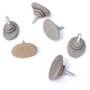 Może przedstawiać: Zestaw sześciu kamiennych gałek z srebrnymi śrubami. Gałki wykonane są z gładkich, szarych i beżowych kamieni rzecznych, niektóre ułożone w stosy. Śruby ułatwiają montaż na meblach. Wymiary w centymetrach.