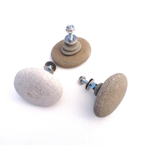 May include: Three natural stone cabinet knobs with metal hardware. The knobs are various shades of gray and brown, with a bolt and washer attachment. One knob is a stack of stones. The knobs are on a white background.