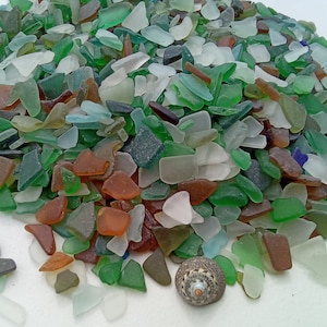 May include: A large pile of colorful sea glass in various shapes and sizes. The sea glass is mostly green, blue, and brown, with some white and clear pieces. There are also two small seashells on the white surface.
