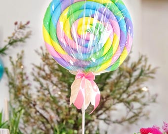 Peut inclure: Une grande décoration en forme de sucette ronde et en spirale, arborant un motif multicolore vif composé de rayures roses, jaunes, bleues et vertes. La sucette est enveloppée dans du plastique transparent et nouée à la base par un ruban rose.