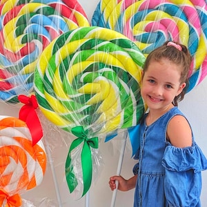 Puede incluir: Grandes accesorios de piruletas redondas con patrones en espiral envueltos en plástico transparente. Una piruleta presenta espirales verdes y amarillas con una cinta verde, otra tiene espirales rojas, azules y amarillas con una cinta roja, y una más pequeña es naranja y blanca.