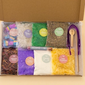 May include: An open cardboard box filled with colorful craft supplies. The box contains bags of rainbow gems, magic crystals, crushed emeralds, unicorn food, precious petals, fairy powder, stardust, and fairy sprinkles. Also included are a wooden spoon, purple tweezers, and a marker.