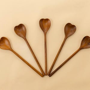 May include: Five wooden spoons with heart-shaped bowls and long, slender handles are arranged in a fan shape. The spoons are made of a warm, medium-toned wood and rest on a light tan surface. The spoons are a kitchen utensil.