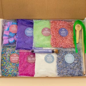 May include: A cardboard box containing various craft supplies in clear plastic bags, including blue and pink cubes, purple beads, green powder, red crystals, multicoloured sprinkles, pink powder, white powder, blue flakes, a glass vial, and green tongs.