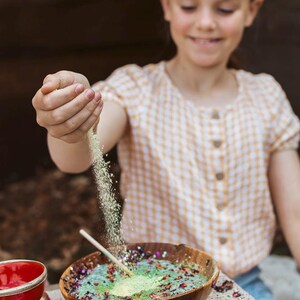 May include: A young person is shown pouring a fine, light-colored powder into a wooden bowl filled with colorful ingredients. The bowl sits on a lace-trimmed surface, with a small red bowl and glass vials nearby. The person wears a yellow and white checkered shirt.