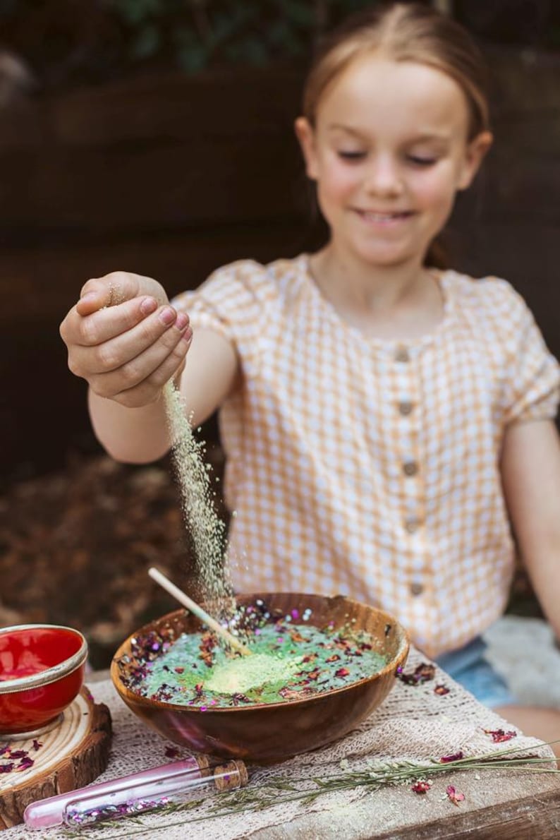 May include: A young person is shown pouring a fine, light-colored powder into a wooden bowl filled with colorful ingredients. The bowl sits on a lace-trimmed surface, with a small red bowl and glass vials nearby. The person wears a yellow and white checkered shirt.