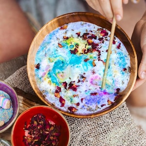 May include: A wooden bowl containing a colourful mixture of blue, purple, and white, decorated with rose petals and herbs. A small red bowl holds more rose petals. A wooden stick stirs the mixture.
