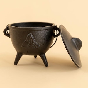 May include: A black cast iron cauldron with three legs and a lid. The cauldron features two handles and a Celtic knot design. The lid is open, showing the inside. A decorative item.