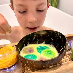 May include: A child gazes at a coconut shell bowl containing colourful bath bombs in a bubbly yellow liquid. A jar of orange bath salts sits beside the bowl on a wooden tray. The bath bombs are green and purple.
