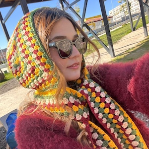 May include: A colorful crochet hood and scarf set in shades of yellow, green, red, and cream. The hood is worn over the head, and the scarf is draped around the neck. The person is wearing sunglasses and a burgundy sweater.