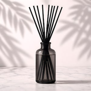 May include: A dark gray glass diffuser bottle filled with liquid and black reeds. The bottle sits on a white marble surface, with shadows of leaves in the background. The reeds are arranged to fan out from the bottle.